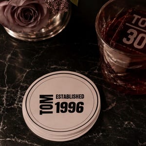 May include: A round, off-white coaster featuring a black circular border and the printed text "TOM ESTABLISHED 1996" in a bold, sans-serif font. The coaster is positioned next to a glass containing a large ice cube with the text "TO 30".