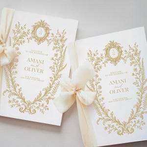 Peut inclure: Deux programmes de mariage ivoire avec des motifs floraux dorés et les noms "AMANI and OLIVER". Chaque programme est noué avec un ruban et un nœud crème. Le texte "WELCOME TO THE WEDDING OF" est visible.