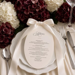 May include: An oval-shaped paper menu with black text and a cream-coloured satin ribbon bow tied at the top, resting on a white scalloped dinner plate. The setting includes a silver spoon, a silver fork, a silver knife, and a clear glass wine goblet.