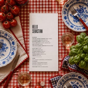 May include: A white menu card featuring the text HELLO SEBASTIAN and a list of food items, set on a red and white chequered tablecloth. The table includes blue and white patterned plates, cutlery, fresh tomatoes, and a potted basil plant.