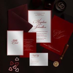 May include: A wedding invitation suite in burgundy and silver, featuring an invitation, RSVP card, and details card. The invitation includes the names "Angelina and Johnathon". Gold wedding rings and a ring box are also present.