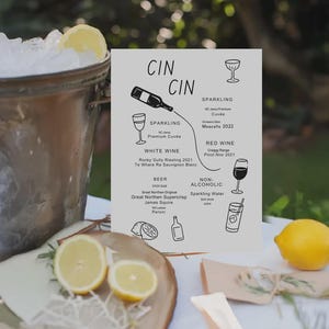 Puede incluir: Escena al aire libre con un cubo de hielo de metal, una tabla de madera con rodajas de limón y un menú. El menú enumera vinos espumosos, blancos y tintos, cerveza y bebidas sin alcohol. El menú está frente a un fondo verde.