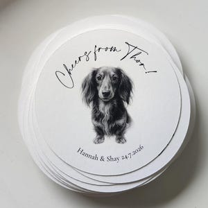 May include: A stack of circular white paper coasters featuring a black and white illustration of a long-haired dachshund. The text "Cheers from Thor!" is printed above the dog, and "Hannah & Shay 24.7.2026" is printed below the illustration.