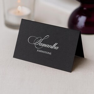 May include: A black place card with the name "Samantha" in elegant white script and the last name "Johnstone" in a smaller, simple font. A lit candle in a glass holder and a dark purple glass are in the background.