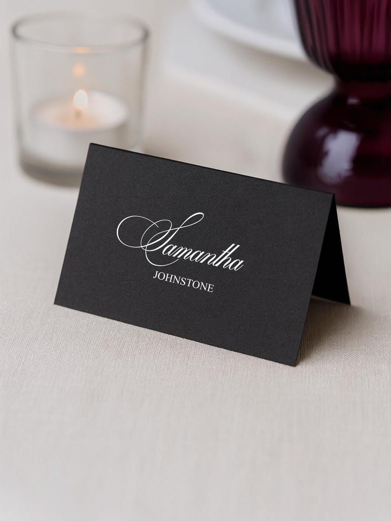 May include: A black place card with the name "Samantha" in elegant white script and the last name "Johnstone" in a smaller, simple font. A lit candle in a glass holder and a dark purple glass are in the background.
