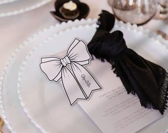May include: A white place card with a black and white bow design. The place card has the name "Chantelle" written on it. The place card is on a white plate with a beaded edge.