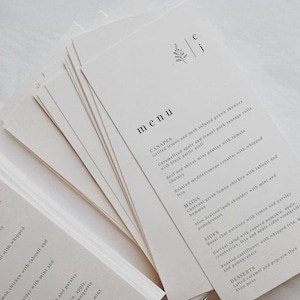 May include: A pile of cream-coloured menu cards, each printed with a minimalist design. The word "menu" is displayed prominently, with a list of dishes below. A small floral motif and initials are at the top. Ideal for a wedding.