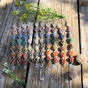 May include: A selection of gemstone chakra pendants in a spectrum of colours, each held in a knotted net. The pendants are arranged in vertical rows and hang against a rustic wooden background. Small charms and a clear quartz crystal are also present.