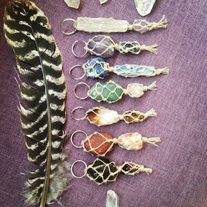Puede incluir: Un conjunto de ocho llaveros de cristal, cada uno con un cristal diferente envuelto en macramé. Los cristales incluyen amatista, lapislázuli, aventurina, cuarzo rosa, ojo de tigre, jaspe rojo, obsidiana negra y cuarzo transparente. Los llaveros se muestran sobre un fondo morado con una pluma.