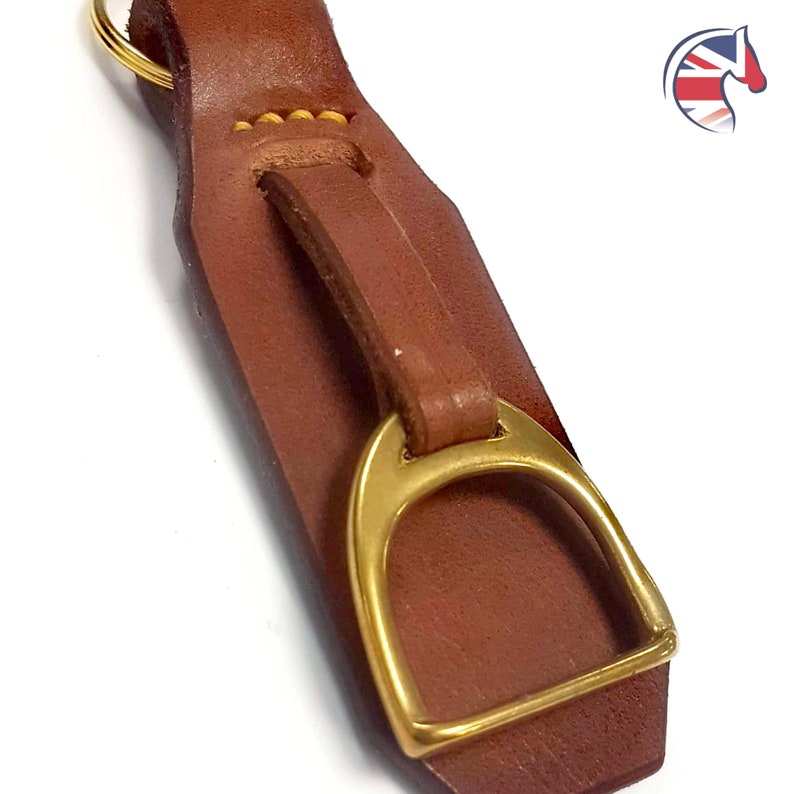 May include: A brown leather keyring featuring a brass stirrup charm attached with a leather loop and yellow stitching. The keyring includes a gold-toned metal ring at the top for attachment. The design is simple, functional, and durable.