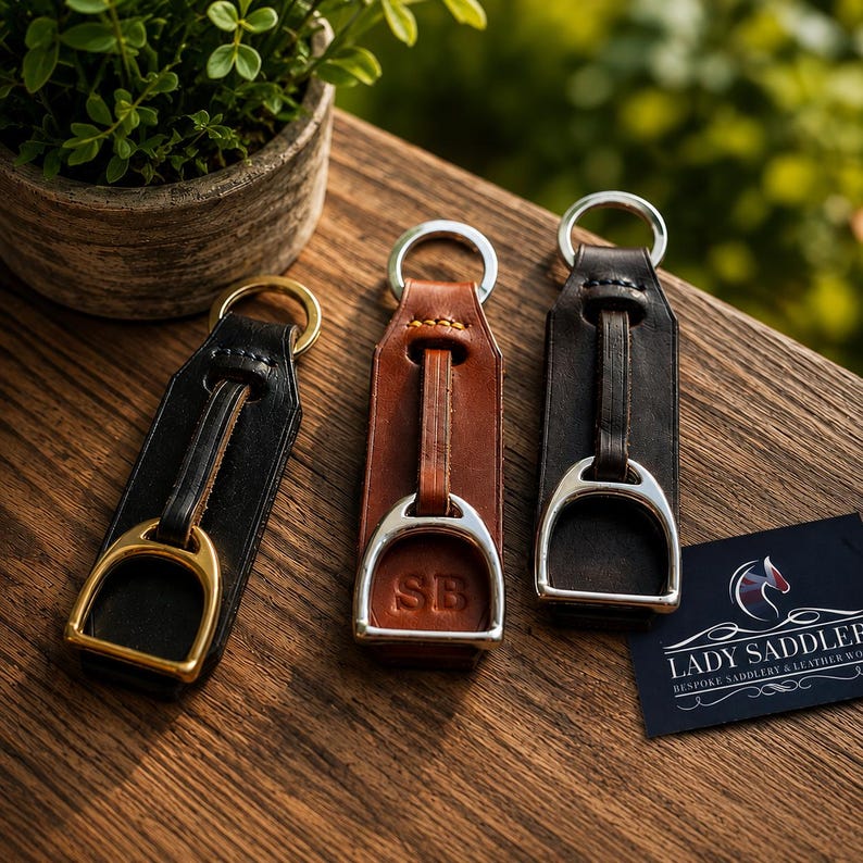 May include: Three leather keychains featuring miniature metal stirrups attached to silver-toned rings. The keychains are black, tan, and dark brown. The tan leather keychain is embossed with the letters SB. A black card displays the text LADY SADDLER.