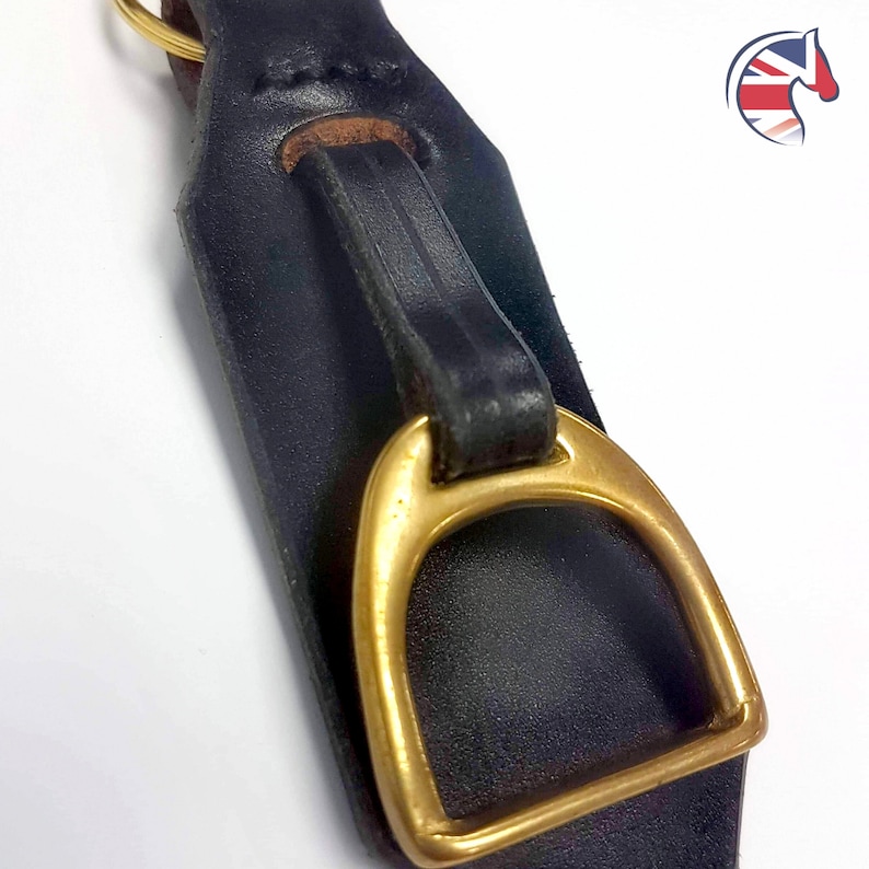 May include: A black leather strap featuring a polished brass stirrup-shaped hardware piece attached with a matching leather loop. The strap has visible stitching along the top edge and a gold-toned metal ring at the very top of the assembly.