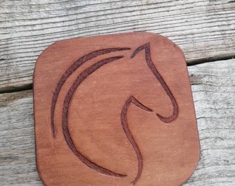 May include: A square-shaped brown leather coaster with rounded corners featuring a debossed, stylised outline of a horse head profile centred on the surface. The leather has a smooth, matte finish with visible natural grain and texture.