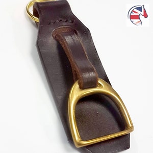 May include: A dark brown leather keyring featuring a polished brass stirrup accent attached with a matching leather loop. The top of the leather strap includes a circular gold-toned metal ring for keys, providing a functional and equestrian-inspired design.