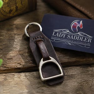 May include: A dark brown leather keyring featuring a silver-toned metal stirrup attachment and a circular split ring. Next to the keyring is a dark blue rectangular business card with the text LADY SADDLER BESPOKE SADDLERY & LEATHER WORK.