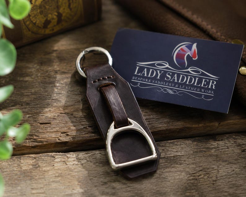 May include: A dark brown leather keyring featuring a silver-toned metal stirrup attachment and a circular split ring. Next to the keyring is a dark blue rectangular business card with the text LADY SADDLER BESPOKE SADDLERY & LEATHER WORK.