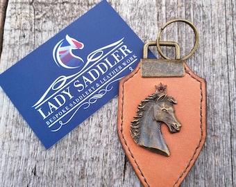 May include: A tan leather keyring shaped like a shield with a bronze-coloured horse head emblem and a metal ring. Next to it is a blue rectangular card with the white text LADY SADDLER BESPOKE SADDLERY & LEATHER WORK and a horse head logo.