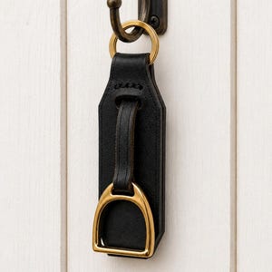 May include: A black leather keyring featuring a gold-toned metal stirrup accent attached to a matching gold-toned ring. The keyring is suspended from a dark bronze-coloured metal wall hook, showcasing a structured design with visible stitching.