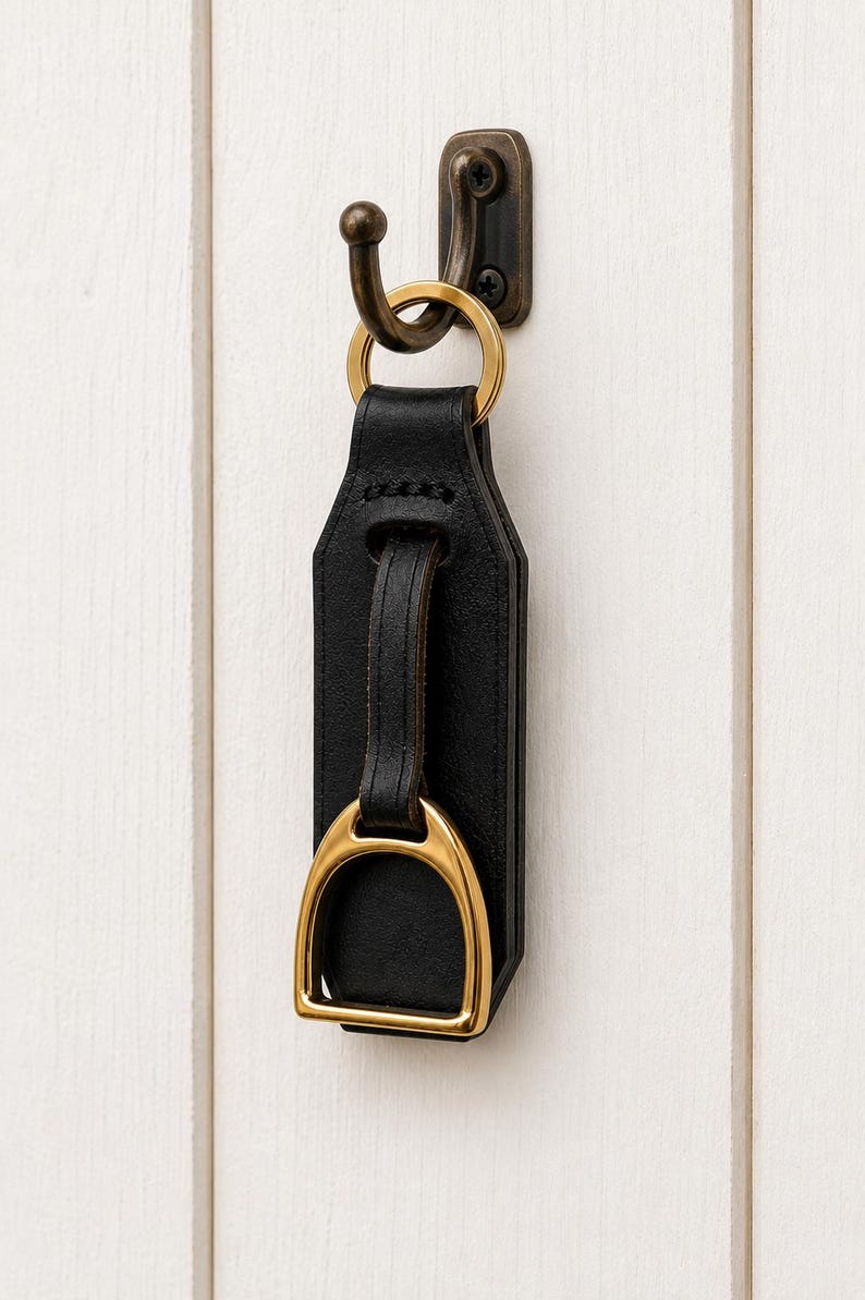 May include: A black leather keyring featuring a gold-toned metal stirrup accent attached to a matching gold-toned ring. The keyring is suspended from a dark bronze-coloured metal wall hook, showcasing a structured design with visible stitching.
