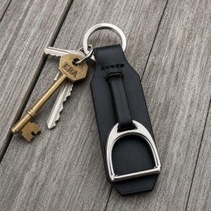 May include: A black leather keyring featuring a silver-toned metal stirrup attachment and a circular split ring. Attached to the ring is a brass-coloured metal key with the word ERA embossed on the head and a standard silver-coloured metal key.