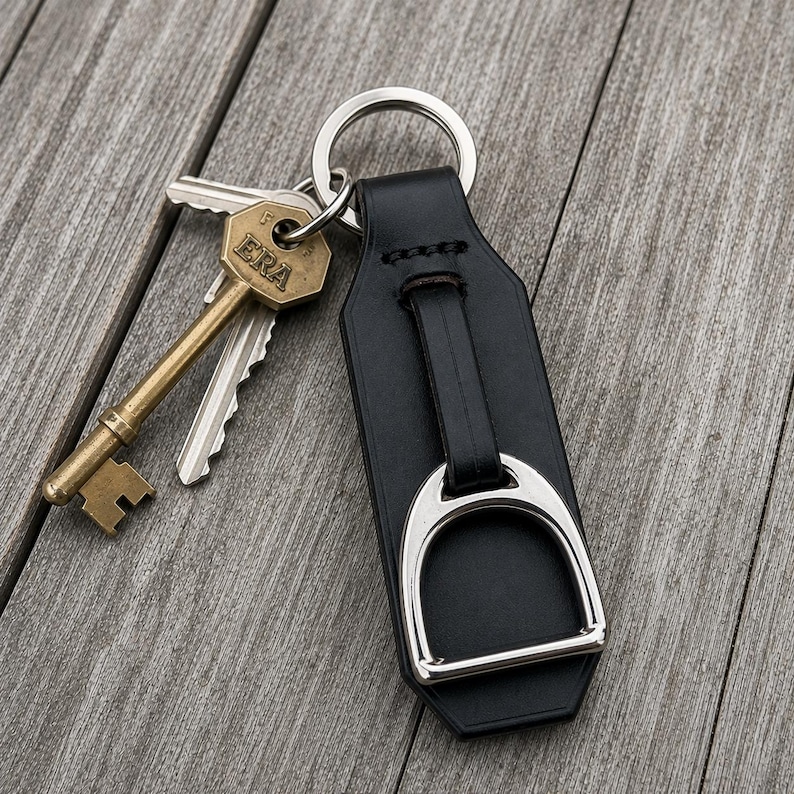 May include: A black leather keyring featuring a silver-toned metal stirrup attachment and a circular split ring. Attached to the ring is a brass-coloured metal key with the word ERA embossed on the head and a standard silver-coloured metal key.
