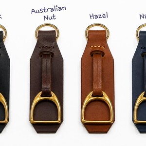 May include: Four leather keychains in black, Australian Nut, hazel, and navy colours. Each keychain features a gold-toned metal stirrup accent attached to a leather strap and a gold-toned circular ring at the top for attachment to keys or bags.