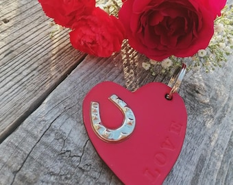May include: A red heart-shaped keyring featuring a silver-toned metal horseshoe emblem attached to the top and the word LOVE embossed vertically along the right side. The keyring includes a circular metal split ring for attachment.