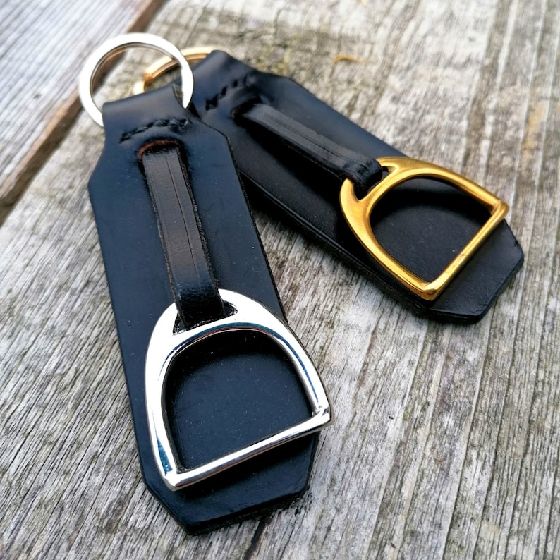 May include: Two black leather keychains featuring a stirrup-shaped metal charm. One keychain has a polished silver-tone stirrup, while the other features a gold-tone stirrup. Each leather strap is secured with a metal ring at the top end.