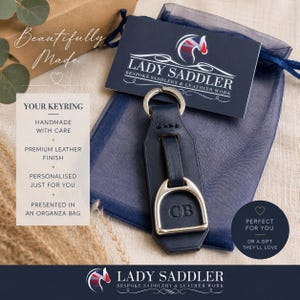 May include: A navy blue leather keyring featuring a silver-toned metal stirrup attachment and a split ring. The leather strap is embossed with the initials CB. The keyring is placed on a dark blue organza bag with a branded card reading LADY SADDLER.