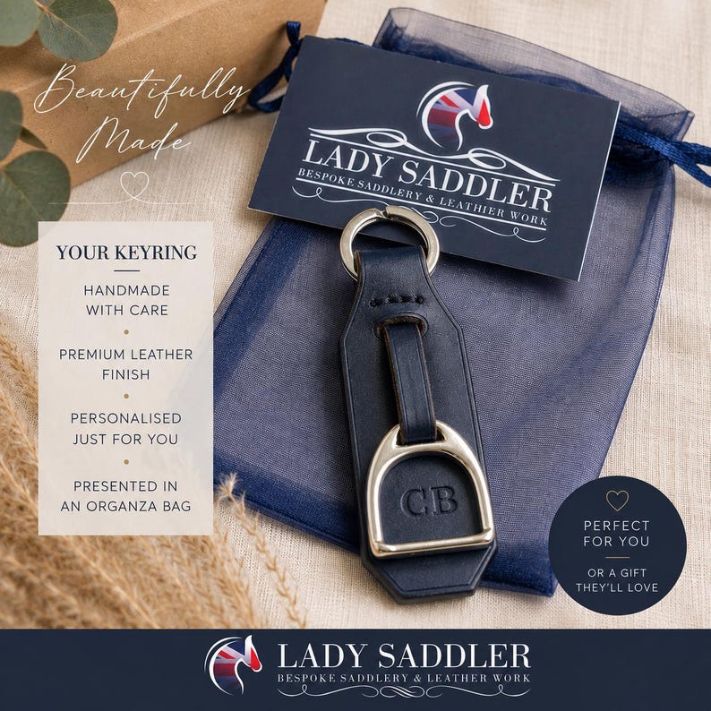 May include: A navy blue leather keyring featuring a silver-toned metal stirrup attachment and a split ring. The leather strap is embossed with the initials CB. The keyring is placed on a dark blue organza bag with a branded card reading LADY SADDLER.