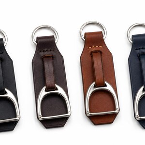 May include: Four leather keyrings in black, dark brown, tan, and navy blue. Each keyring features a silver-toned metal stirrup attached to a leather strap with a matching silver-toned metal split ring at the top for securing keys.