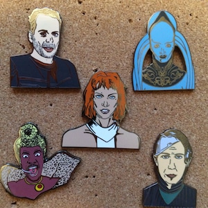 May include: Five enamel pins featuring different characters. The pins are on a corkboard background. The characters include a man with blonde hair, a woman with red hair, a woman with blue skin, a woman with brown skin and a man with brown hair.