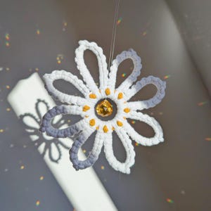 Op de afbeelding: Een handgemaakte gehaakte madelief met witte bloemblaadjes en gele accenten, opgehangen aan een dun draadje. Het midden heeft een sprankelend gele kristal. De schaduw van de bloem is zichtbaar.