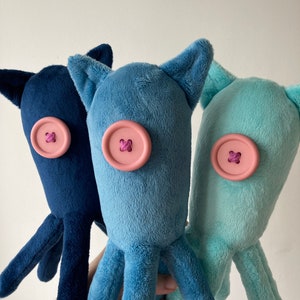 May include: Three plush octopus toys with different colored bodies: navy blue, light blue, and light turquoise. Each octopus has a pink button for an eye with a purple thread cross.