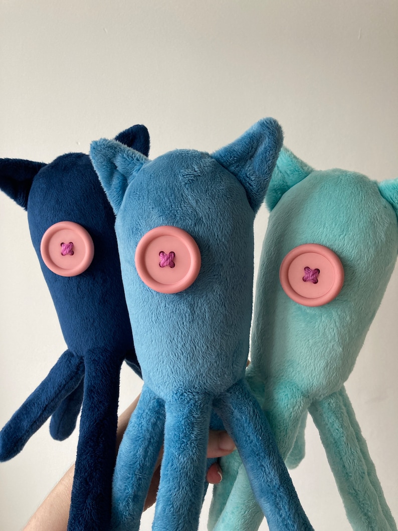 May include: Three plush octopus toys with different colored bodies: navy blue, light blue, and light turquoise. Each octopus has a pink button for an eye with a purple thread cross.