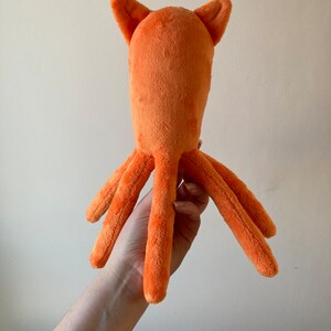 May include: A plush orange octopus toy with a long body and six tentacles. The octopus has two small ears on top of its head.