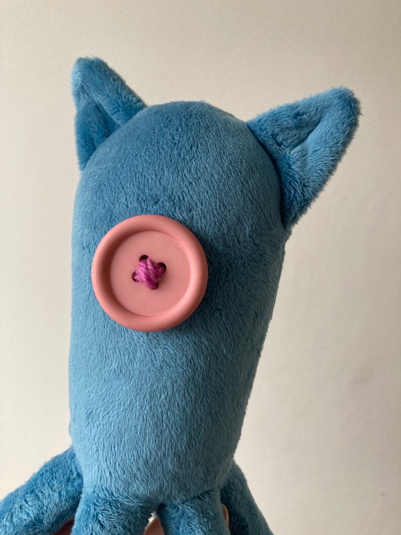May include: A blue plush toy with a pink button for an eye and two floppy ears. The toy has a long body and multiple tentacles.