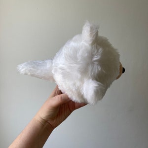 Puede incluir: Juguete de peluche blanco con forma de perro con una cola esponjosa. El juguete se sostiene en una mano con el fondo desenfocado.