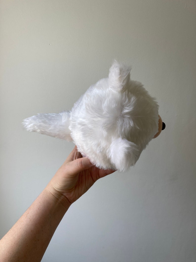 Puede incluir: Juguete de peluche blanco con forma de perro con una cola esponjosa. El juguete se sostiene en una mano con el fondo desenfocado.
