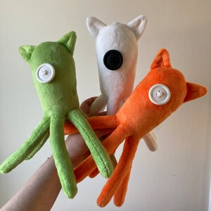 May include: Three plush toys with button eyes. The first toy is green and has eight tentacles. The second toy is white with black button eyes. The third toy is orange with a white button eye.