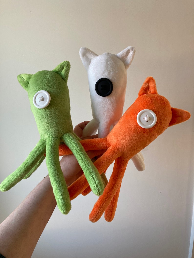 May include: Three plush toys with button eyes. The first toy is green and has eight tentacles. The second toy is white with black button eyes. The third toy is orange with a white button eye.
