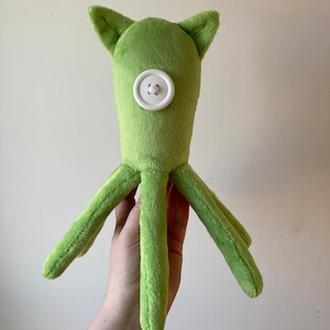 May include: A green plush octopus with a white button for an eye. The octopus has six tentacles.