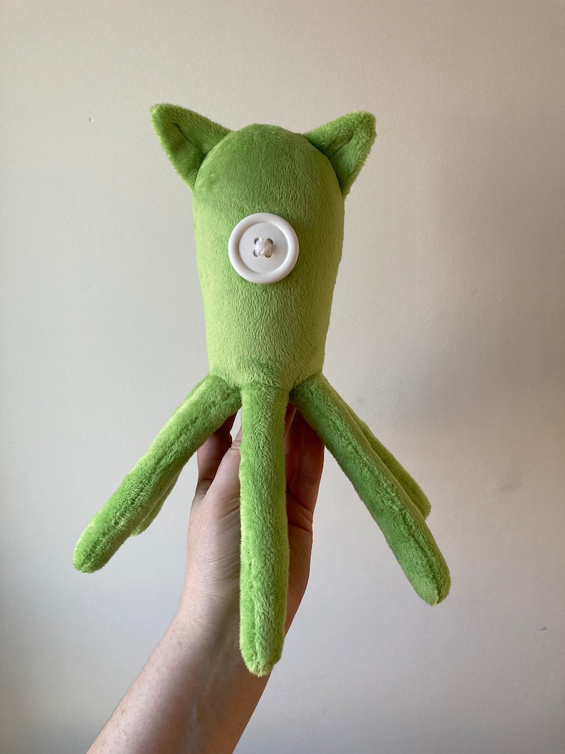 May include: A green plush octopus with a white button for an eye. The octopus has six tentacles.