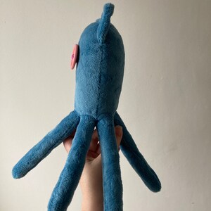 May include: A blue plush octopus toy with a pink button eye. The octopus has six tentacles and is held by a hand.