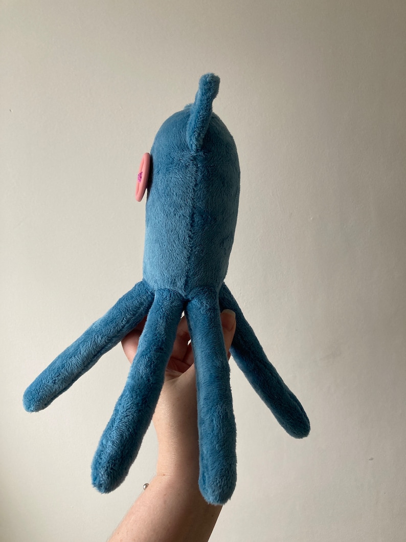 May include: A blue plush octopus toy with a pink button eye. The octopus has six tentacles and is held by a hand.