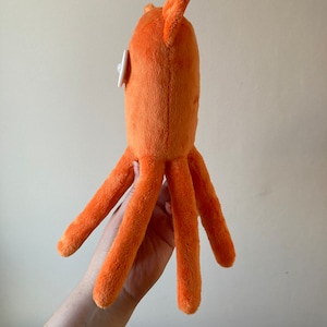 May include: Orange plush octopus toy with white button eyes. The octopus has six tentacles and a rounded body.
