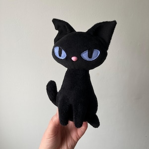 Op de afbeelding: Zwart pluche katten speelgoed met blauwe ogen en een roze neus. De kat zit met zijn oren omhoog.