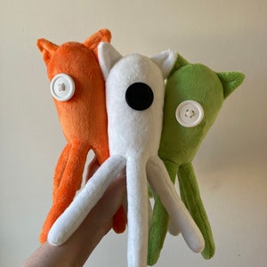 May include: Three plush toys shaped like octopus with different colored bodies. The octopus on the left is orange, the middle octopus is white, and the octopus on the right is green. Each octopus has a button for an eye.