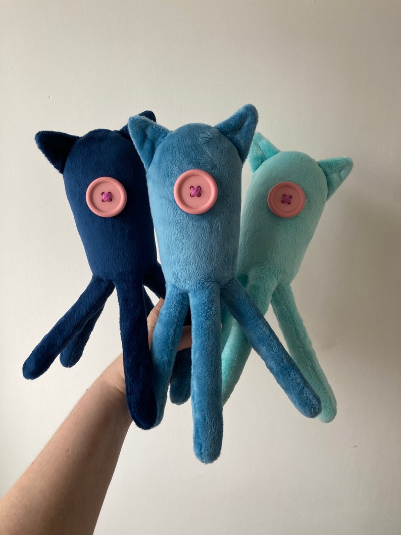 May include: Three plush octopus toys with different colored bodies: navy blue, light blue, and light teal. Each octopus has a pink button sewn on its head with a purple X in the center.