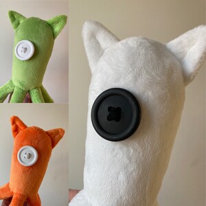 May include: Three plush toys with button eyes. The first is green with a white button, the second is white with a black button, and the third is orange with a white button.